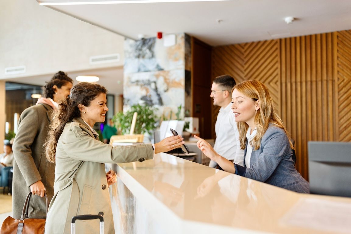 Modern hotel front desk with smiling female guest checking in using a smartphone, assisted by friendly hotel staff.