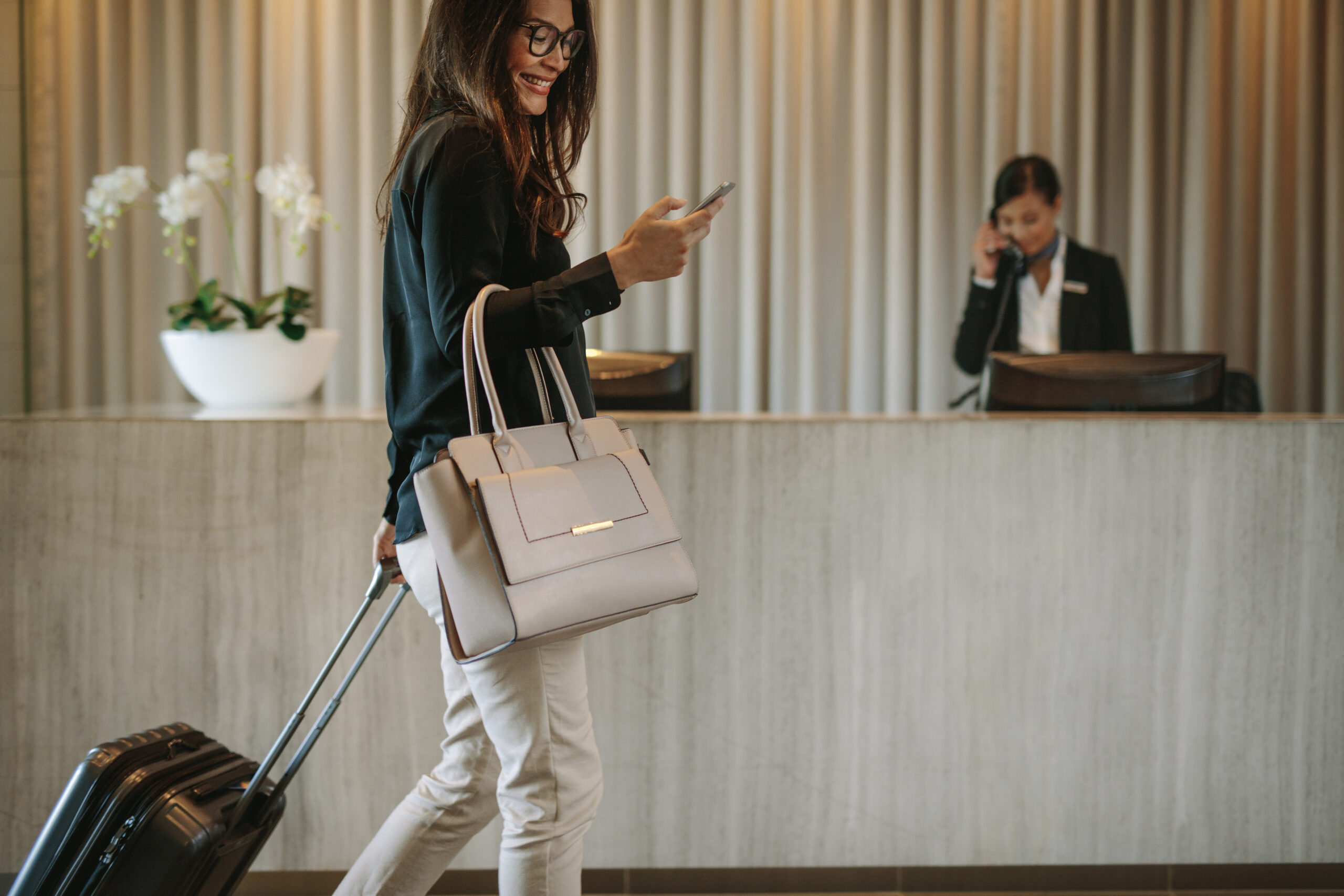 Woman checking into hotel, using her phone, not having to stand in line. Pushing suitcase, while digitally checking in.